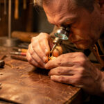 Un bijoutier examine une bague ancienne en or à l’aide d’une loupe sur un établi de maroquinerie éclairé par une lumière chaude.