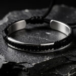 Bracelet menottes en argent poli monté sur un cordon noir et posé sur une pierre sombre, éclairé par une lumière d’atelier.